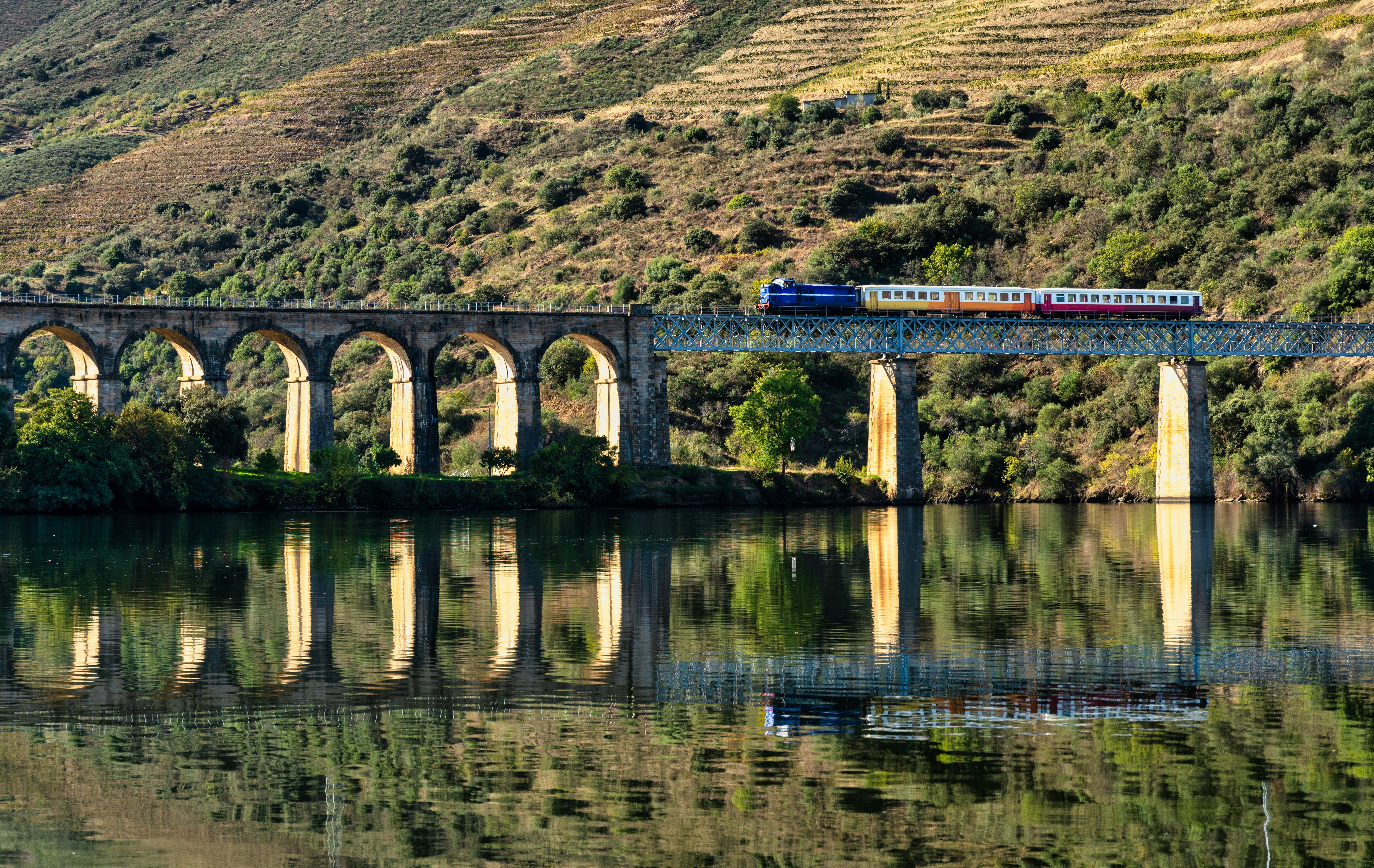 A train on a bridge crossing a river, with the hillside reflected in the water's surface.