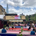 Several diners are sat at colourful benches in the outside area at Flat Iron Square. The Shard can be seen in the background. London's food halls and markets